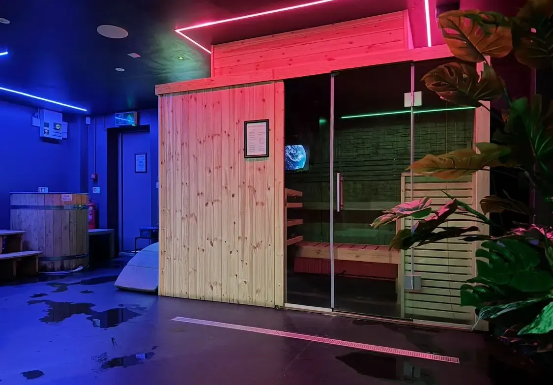 Wooden sauna cabin with glass door and neon-lit contrast therapy suite, wooden cold plunge barrel to the left