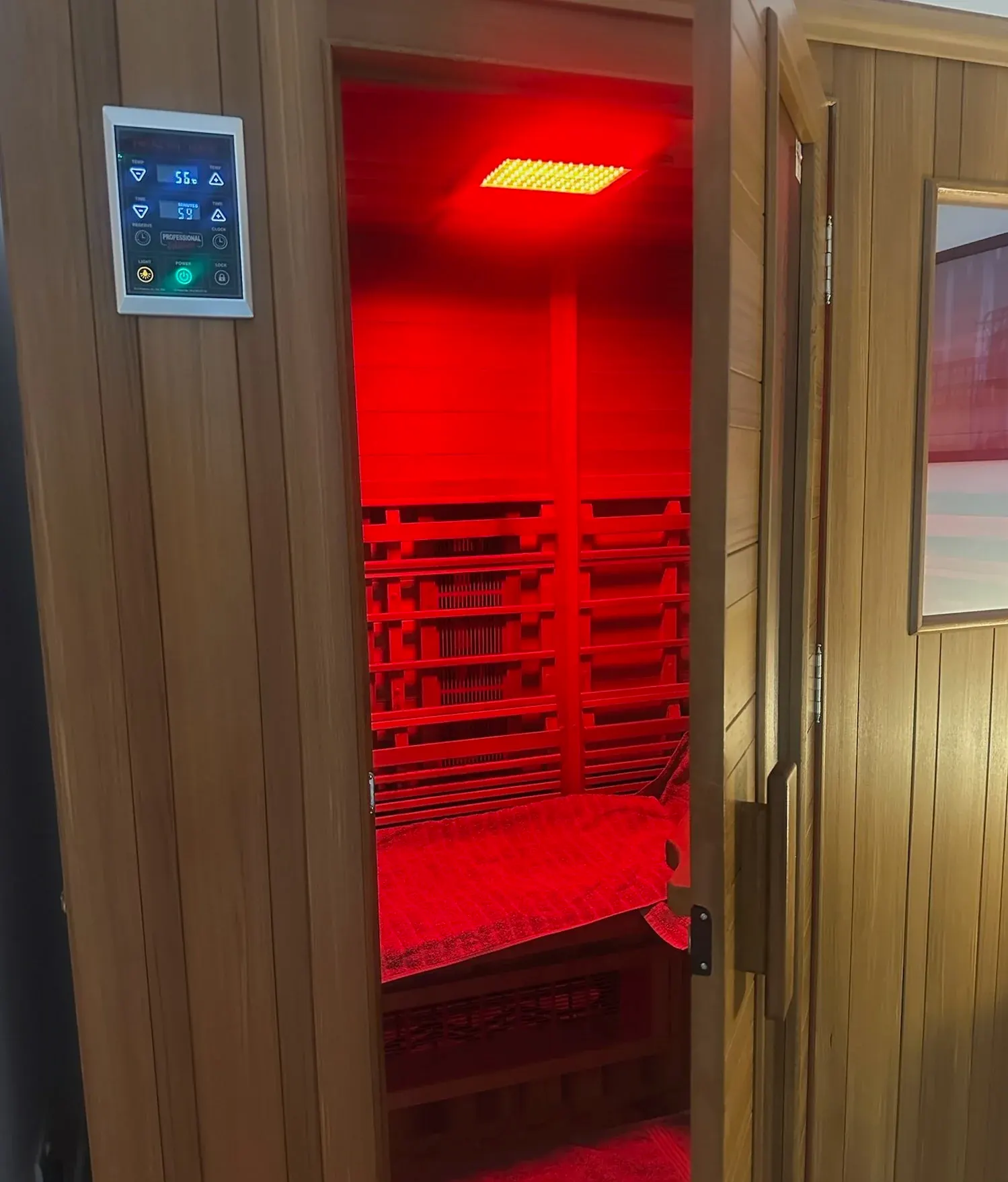 Wooden far infrared sauna cabin with control panel, door open showing red-lit empty interior and bench
