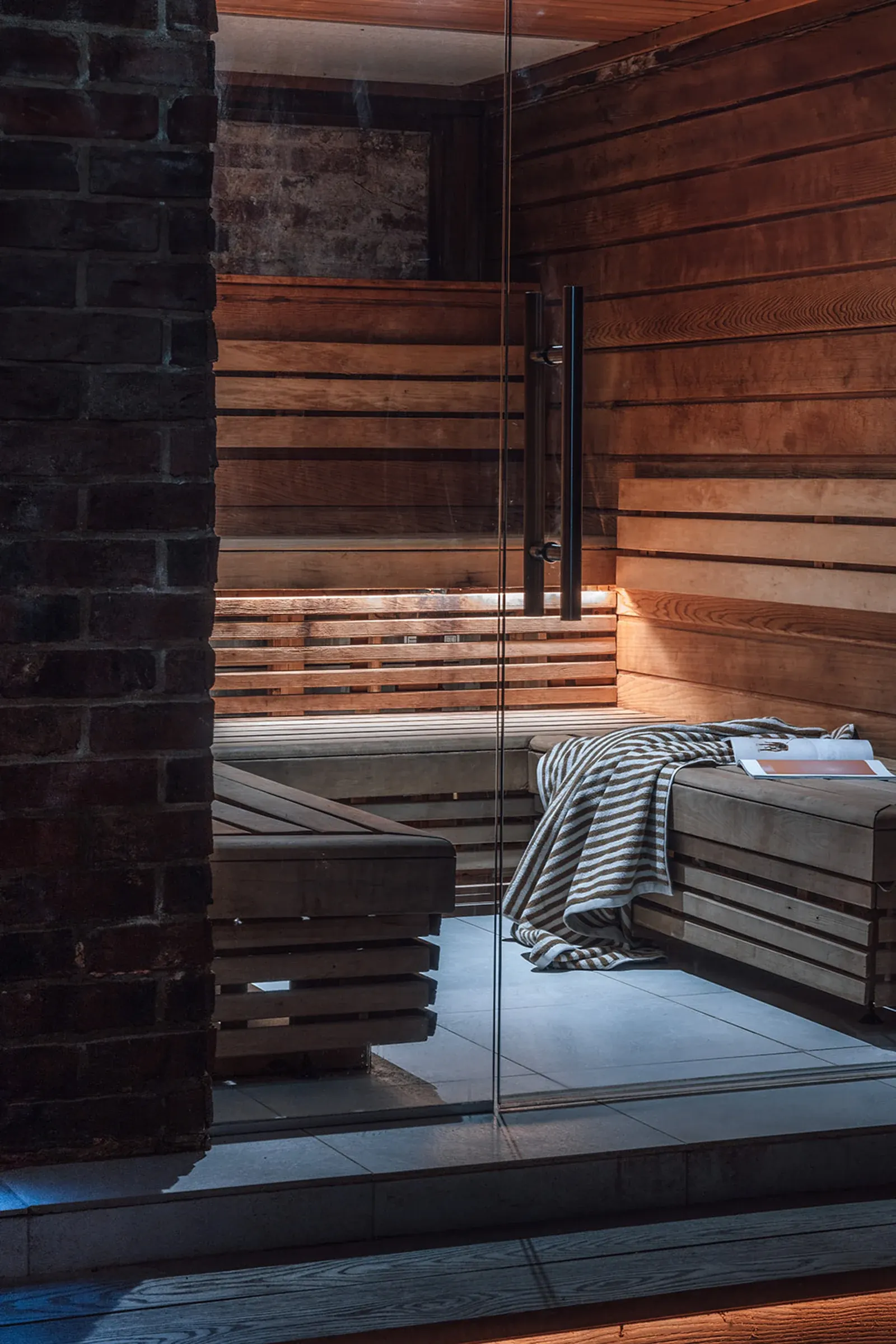 Wood-panelled Scandinavian sauna with tiered benches and a glass door, in the brick-walled spa area, no people