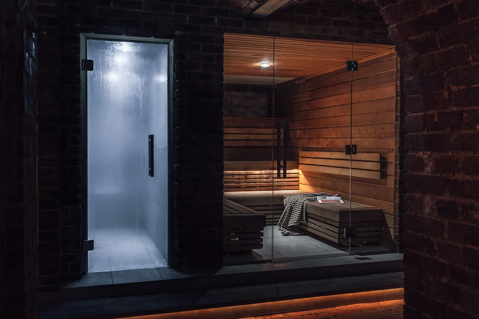 Glass-fronted steam room beside the wood-lined sauna with benches and a throw, set against dark tiling and brickwork, no people