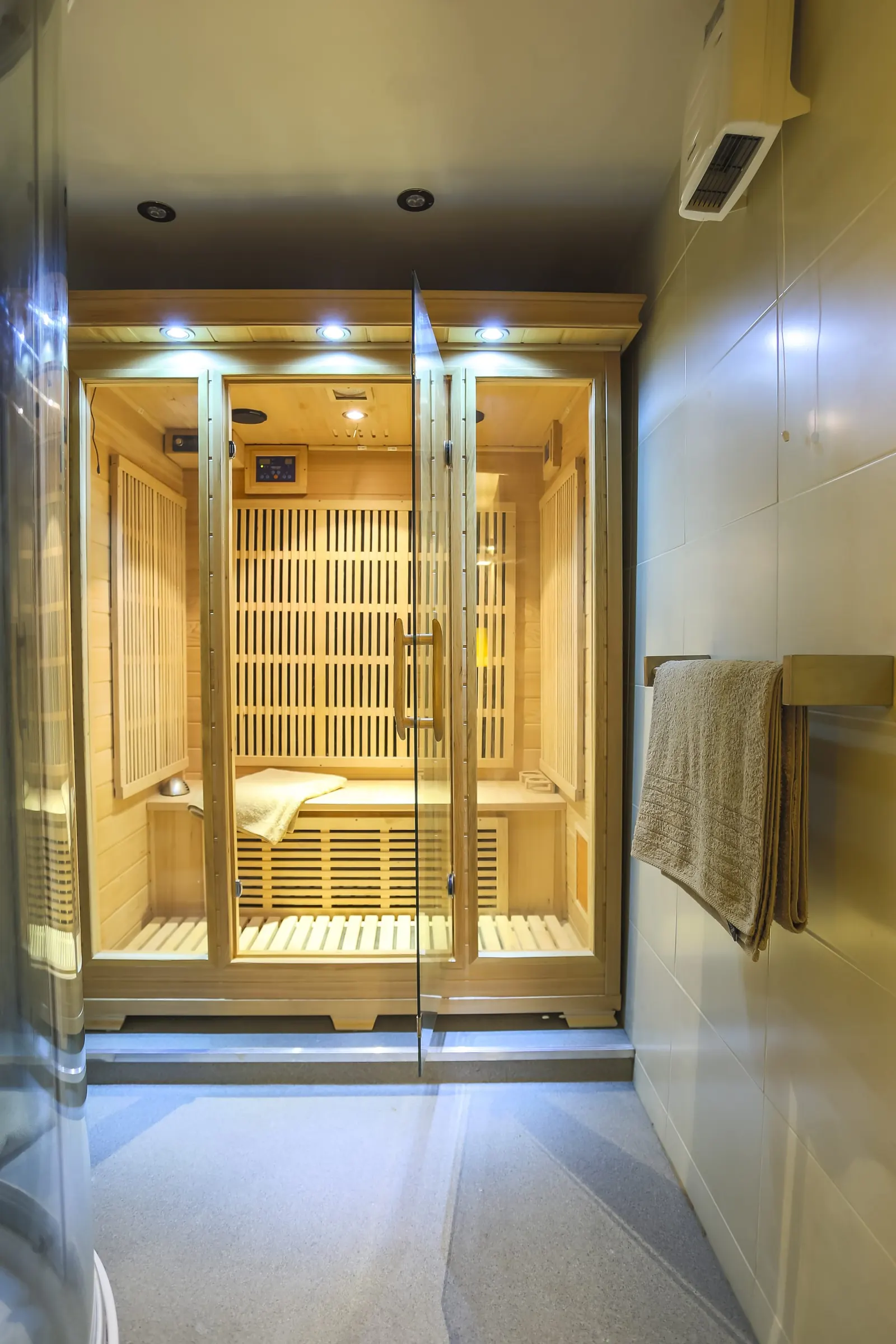 Light-wood far infrared sauna cabin with glass doors, towel on the bench, in a tiled room, no people