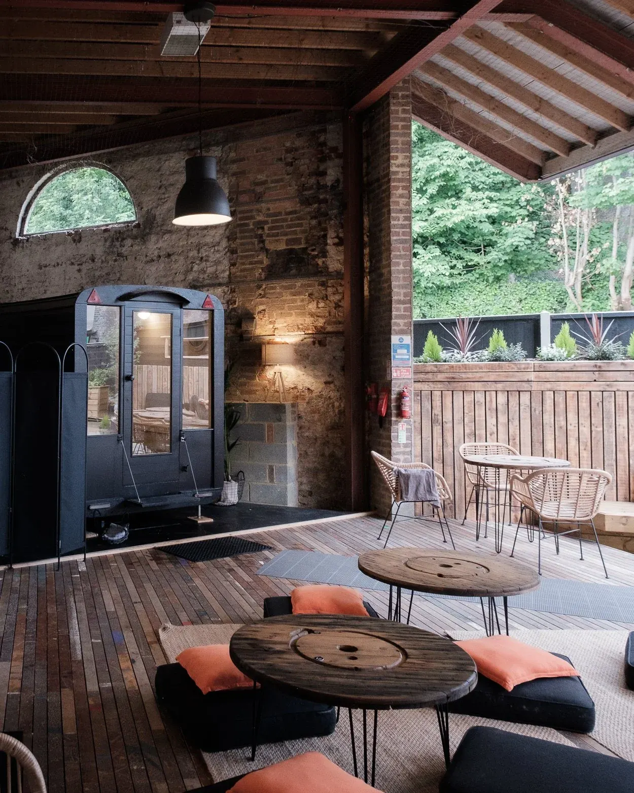 Black shepherd's-hut style sauna cabin with glass door standing on the covered timber deck of the cafe, with low tables and cushions in front