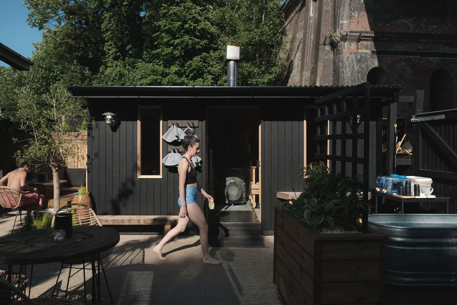Dark-clad sauna cabin and cold-plunge tub on the sunny courtyard under the railway arches, one person walking past