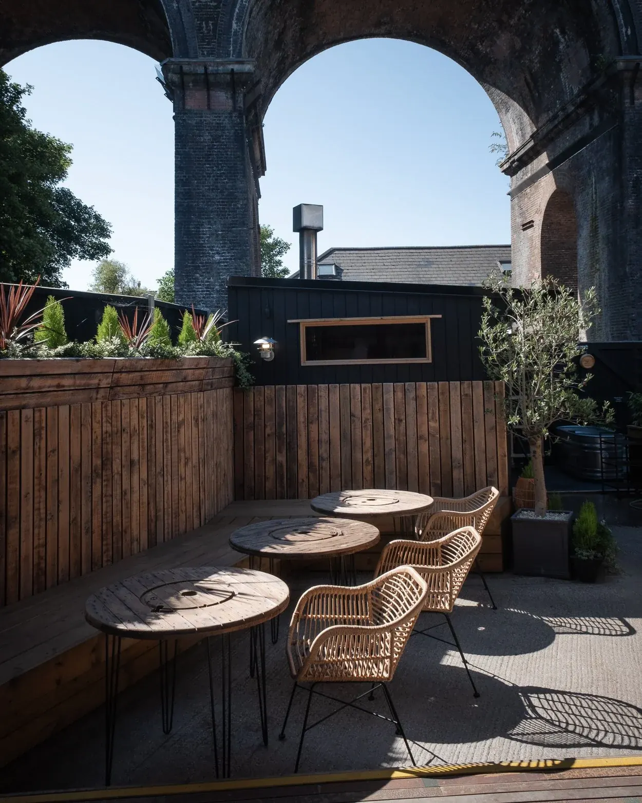 Outdoor terrace with cafe tables under a brick railway viaduct arch, black sauna cabin and plunge tub behind the fence