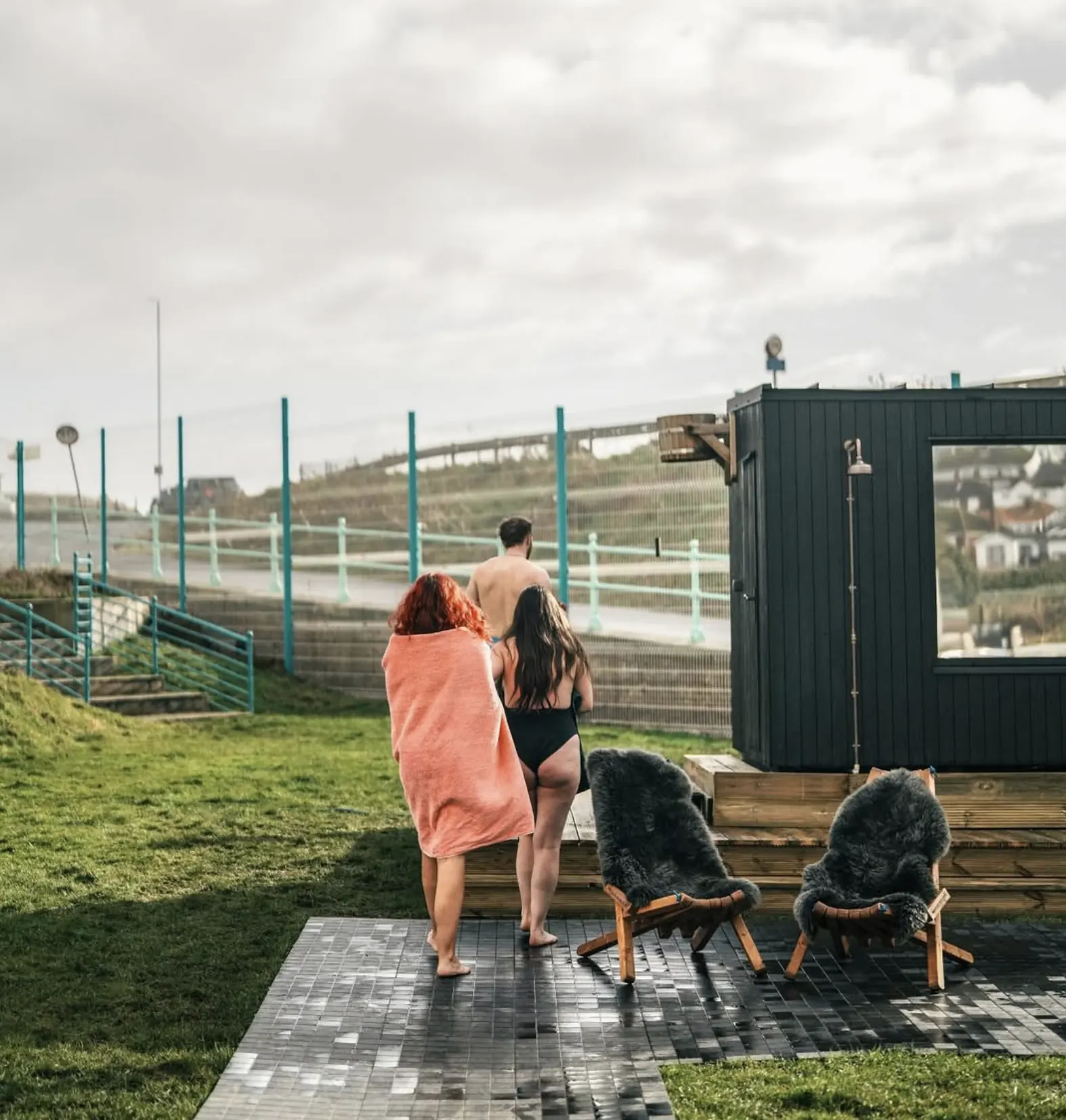 Two guests wrapped in towels walking towards the sauna cabin, grassy hillside behind