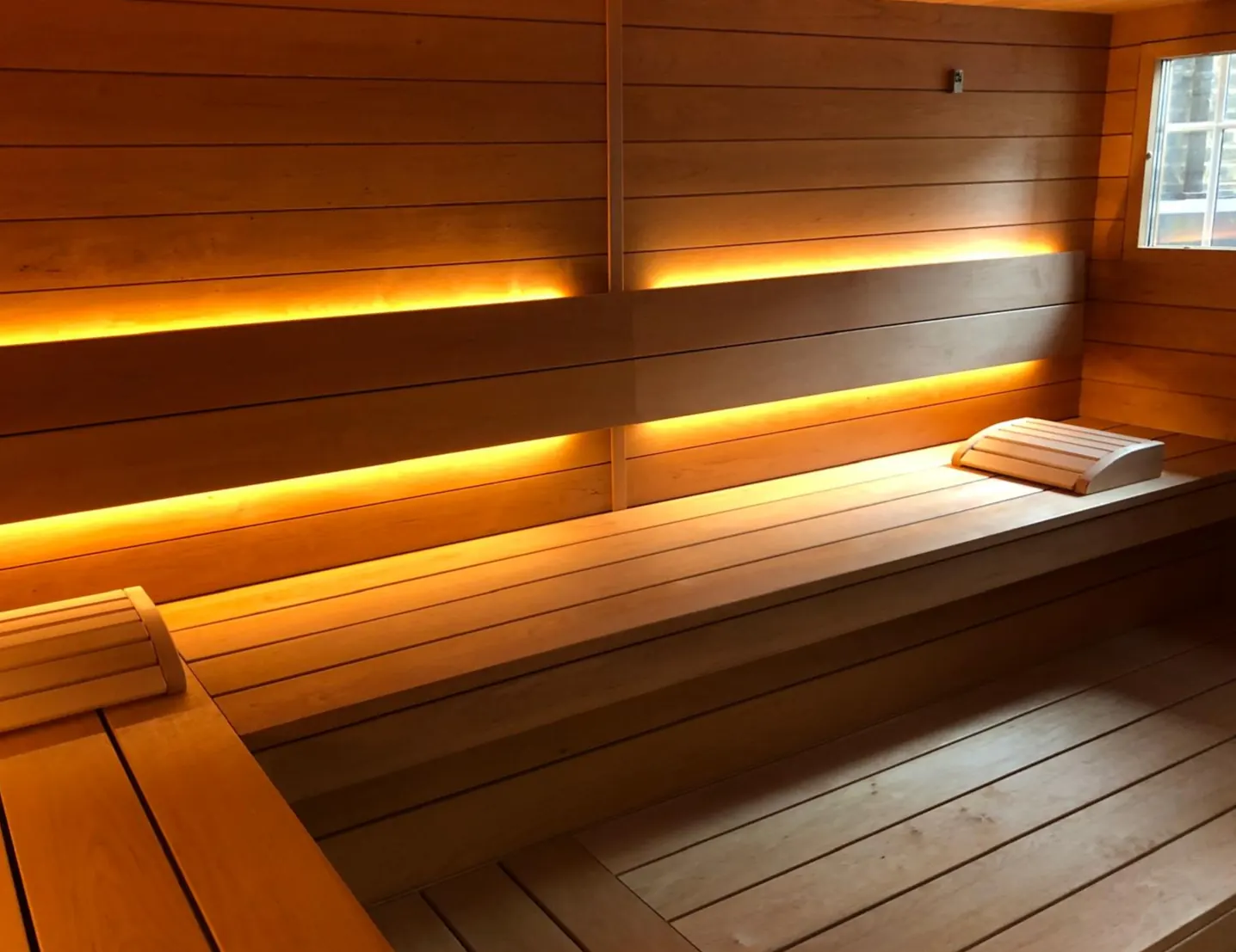 Empty wood-panelled sauna cabin with two tiers of benches, warm LED strip lighting under the benches and a small window