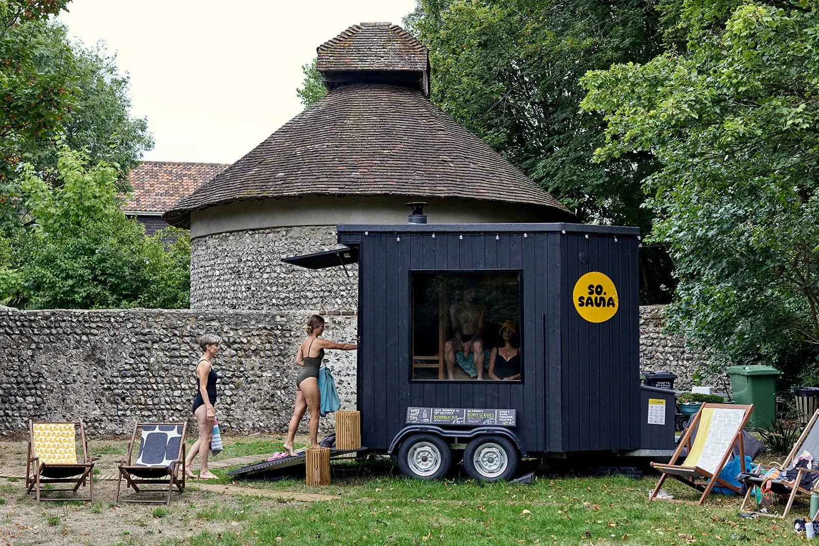 The black horsebox sauna trailer parked on a lawn beside a flint-walled garden, with deckchairs and a few people standing by the ramp